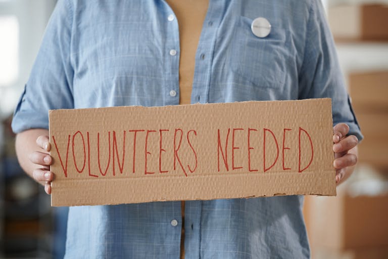 Person holding a sign reading 'Volunteers Needed' to encourage community support.