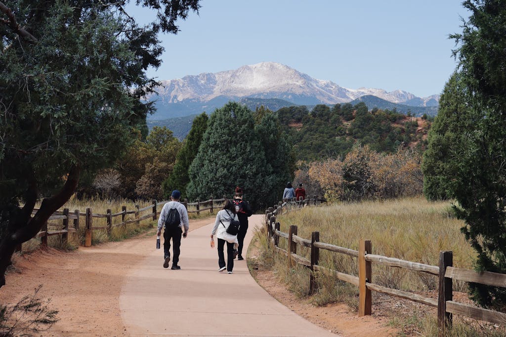 People hiking a scenic trail with wooden fence and mountain backdrop under blue skies.