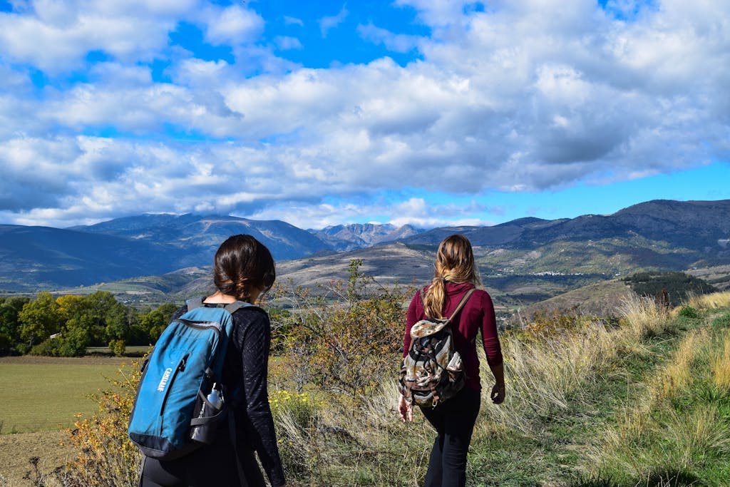 Two women hiking on a mountain trail with scenic views under a blue sky.